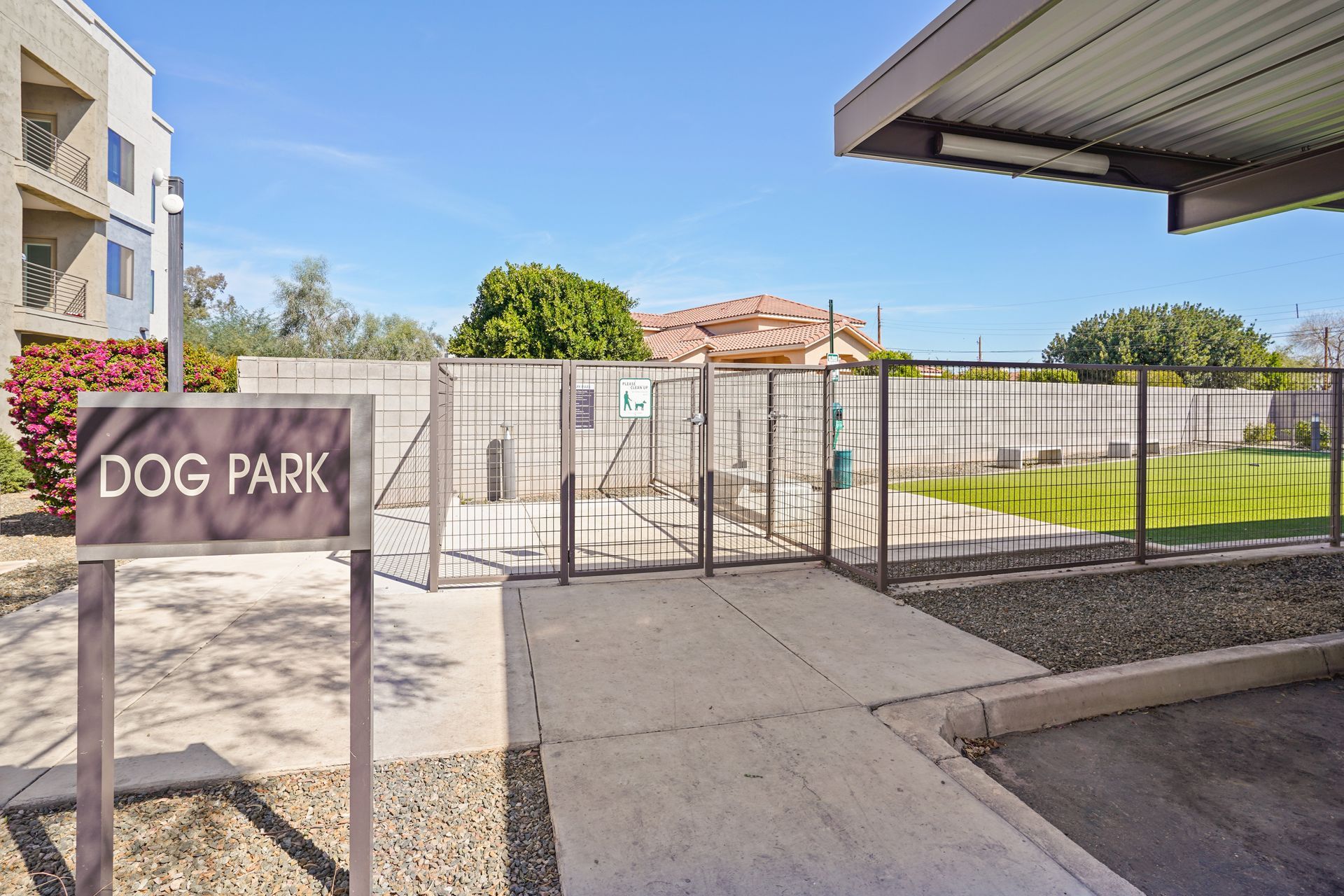 Dog park entrance with sign, fenced area, concrete path, and nearby apartment building at Parc South Mountain in Phoenix, AZ..