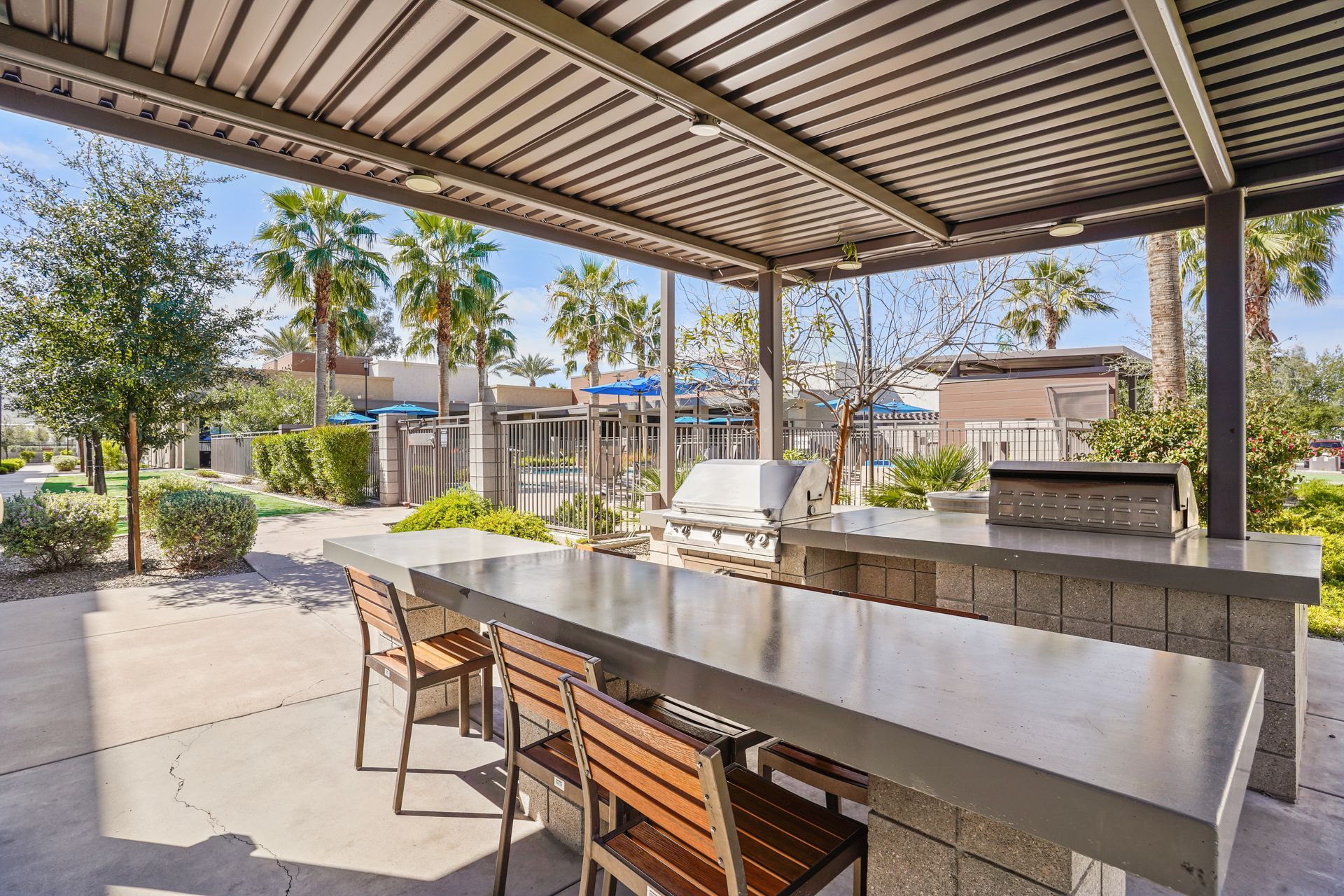 Outdoor kitchen with a grilling area, long concrete counter, bar stools, and overhead shade. Palm trees in background at Parc South Mountain in Phoenix, AZ..