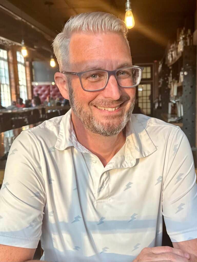 A man with glasses and a beard is smiling while sitting at a table.