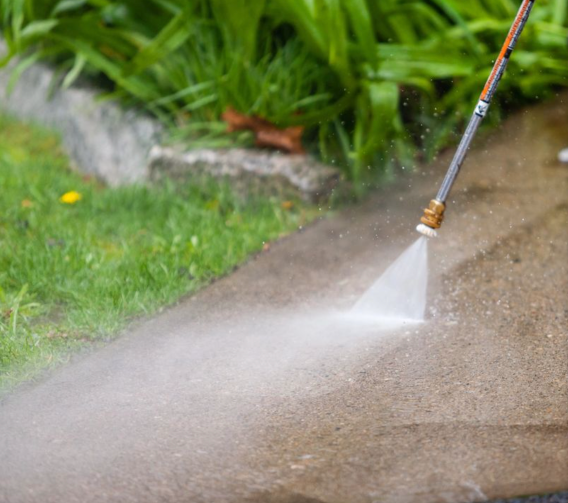 Pressure washer spraying concrete sidewalk; water mist, greenery in the background.