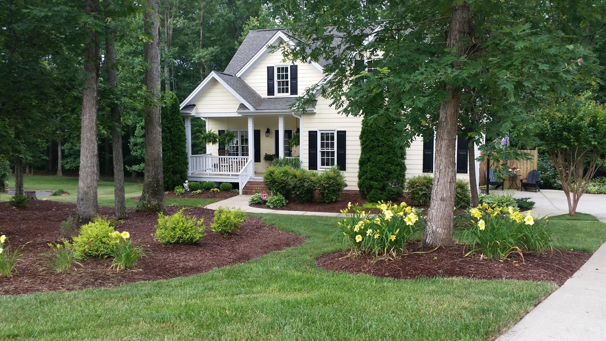 A white house with black shutters is surrounded by trees and flowers.