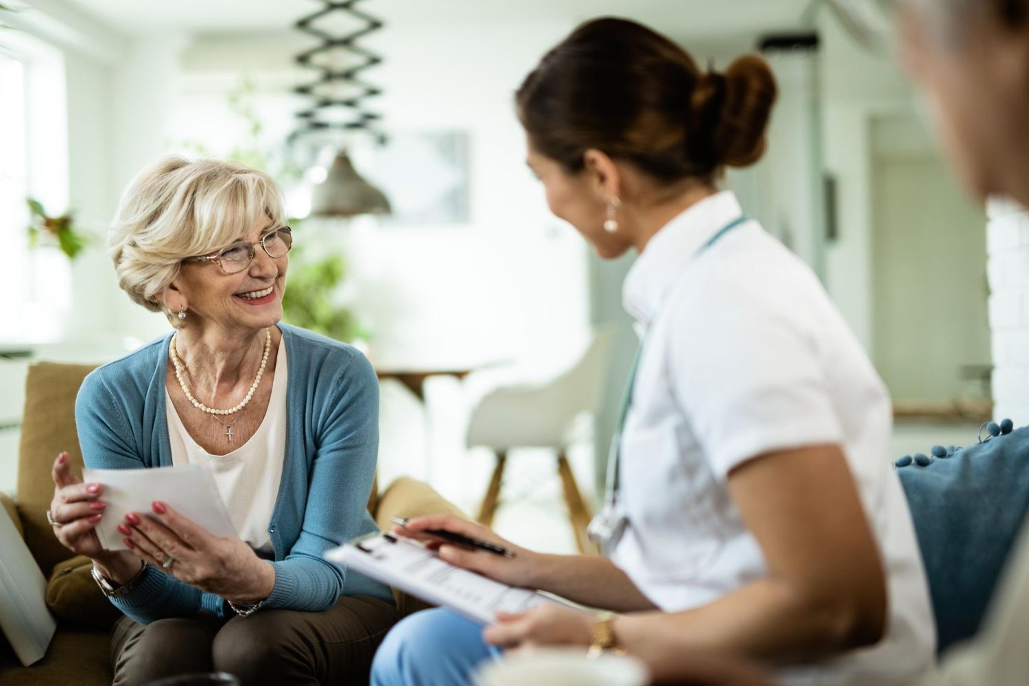 An older woman smiles, holding paperwork, while a healthcare worker takes notes in a living room.