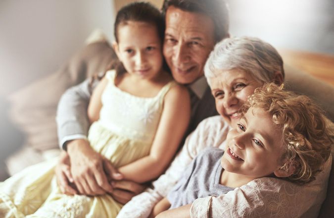 Grandparents hugging two grandchildren on a couch. Smiling, they are inside a home, close together.