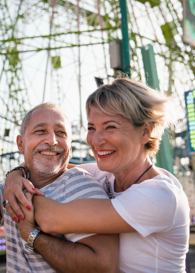 Older couple hugging and smiling at a carnival; Ferris wheel in the background.
