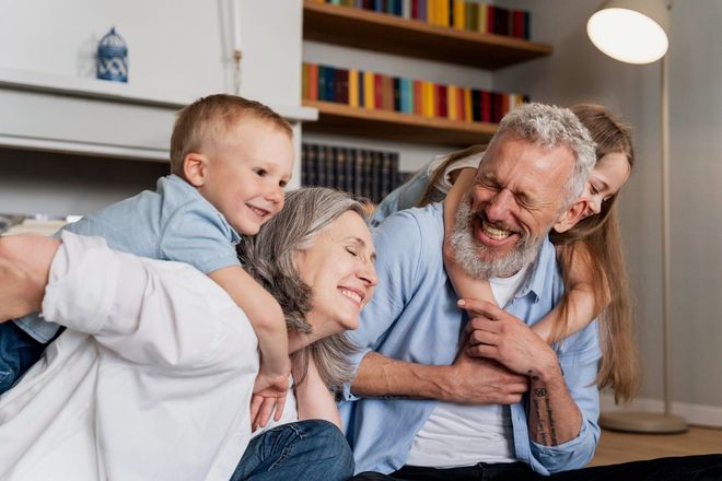 Grandparents laughing with two grandchildren; boy on the woman's back, girl on the man's, indoors.