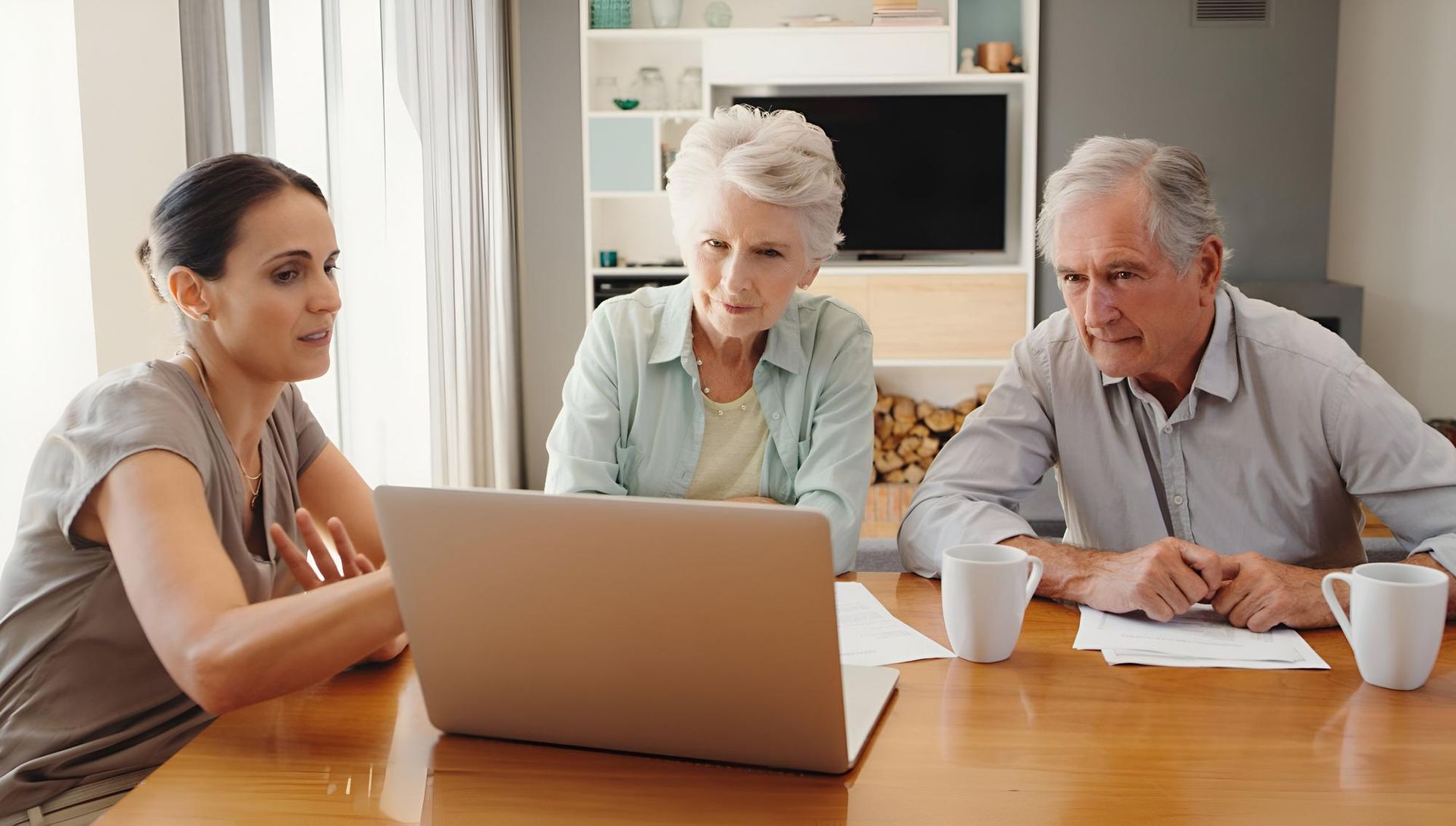 A woman shows a laptop to an elderly couple at a table, likely discussing finances.