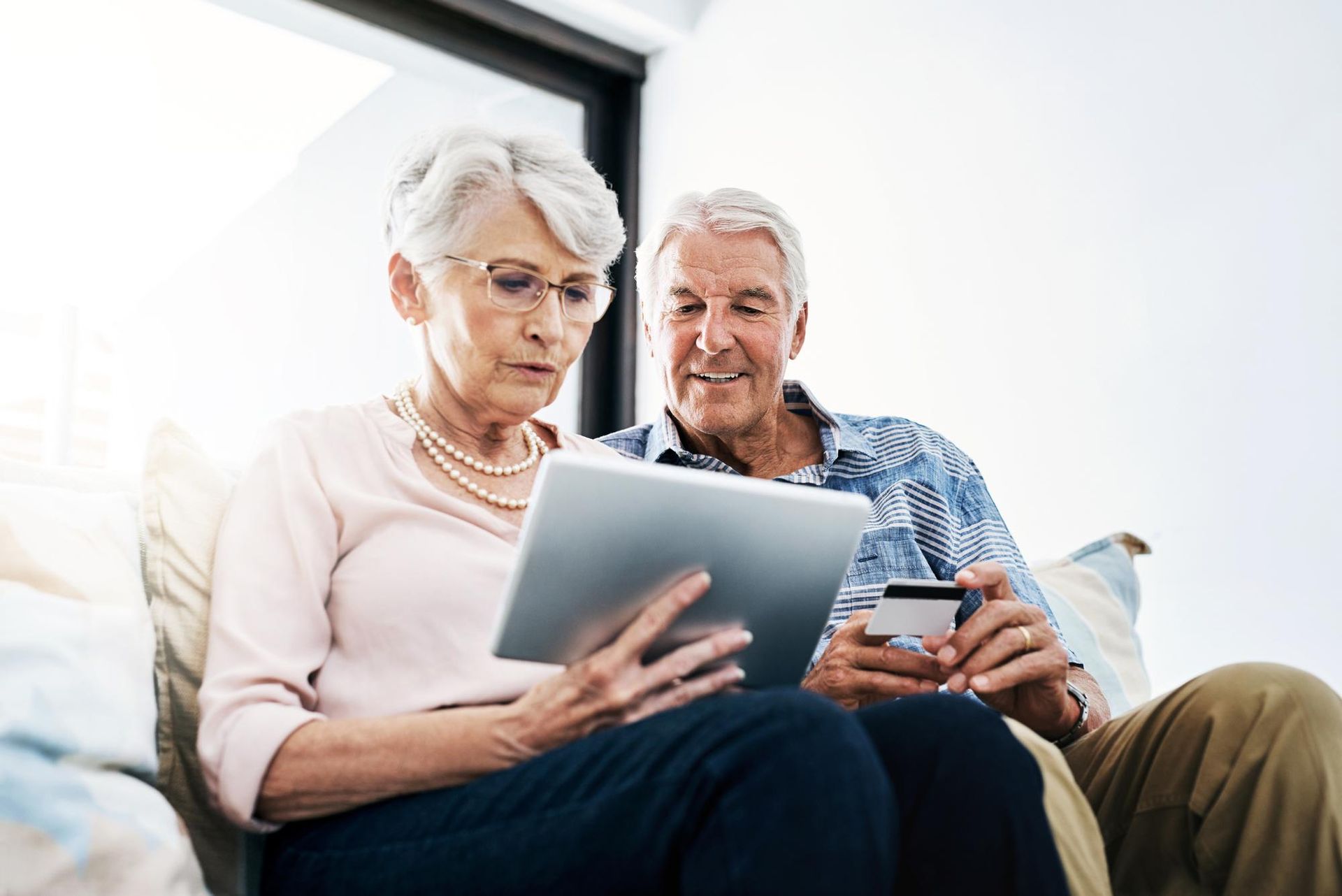 An elderly couple looks at a tablet, the woman holding it while the man holds a credit card.