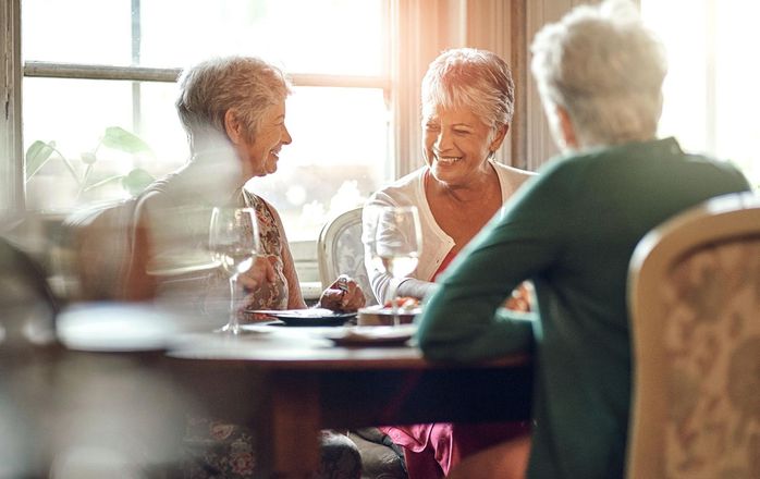 Three older women seated at a dining table, laughing and enjoying a meal with drinks, near a sunlit window.
