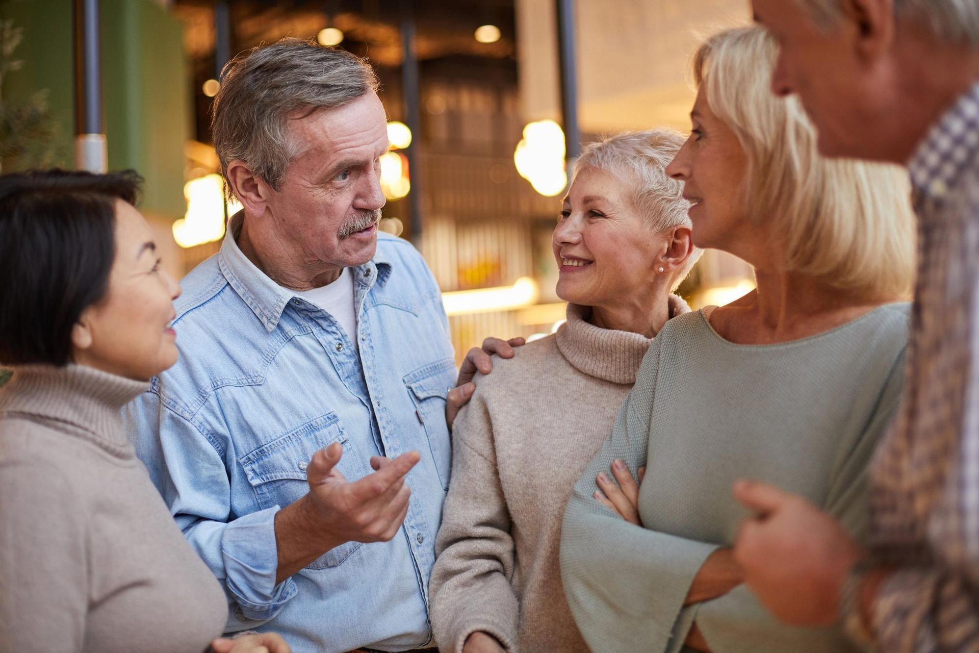 A group of five older adults talking and smiling, indoors. One man gestures, others listen and interact.
