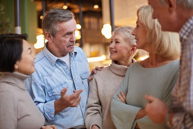 A group of six older adults conversing indoors, with varying expressions and some with arms around each other.