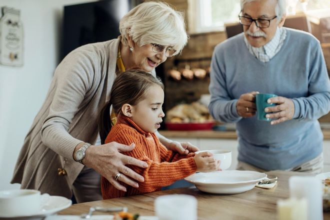 Grandmother helping a young girl at a kitchen table, grandfather holding a mug nearby. They appear to be preparing food.