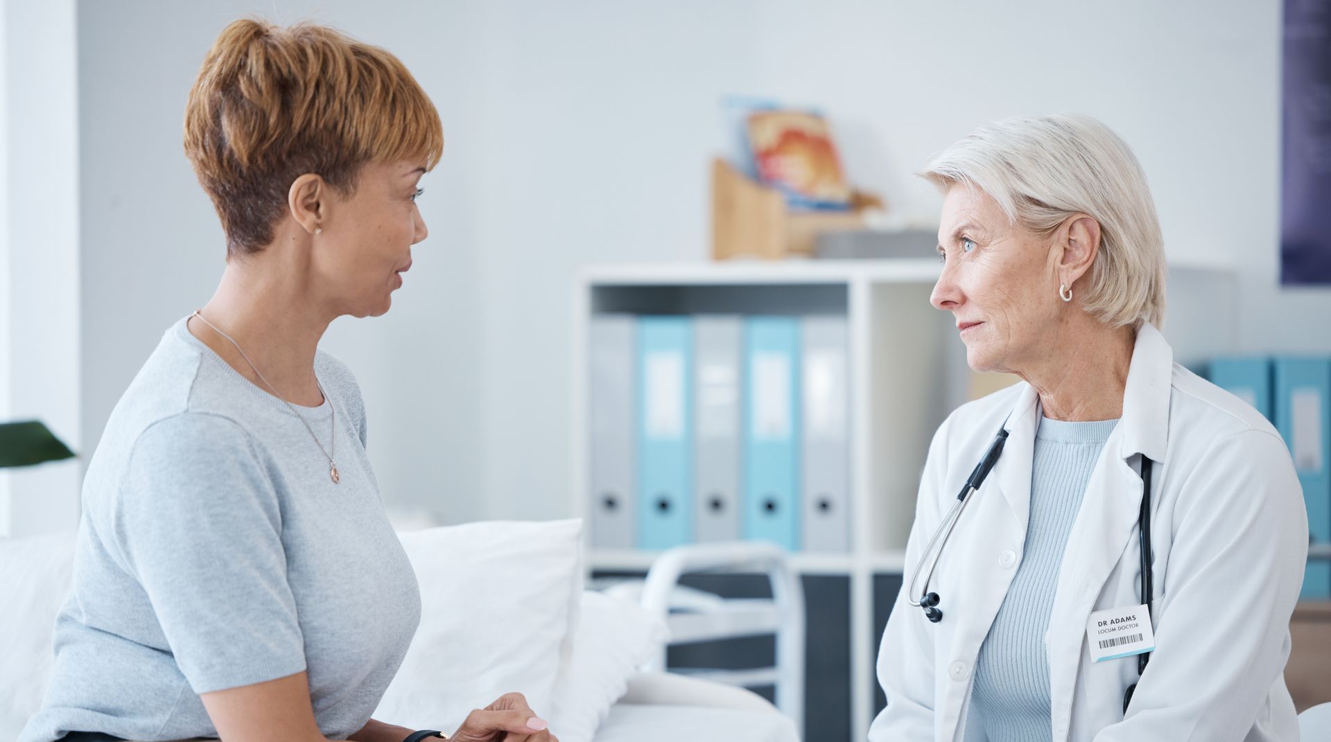 A doctor in a white coat talking to a patient in a light blue top in a bright, well-lit medical office.
