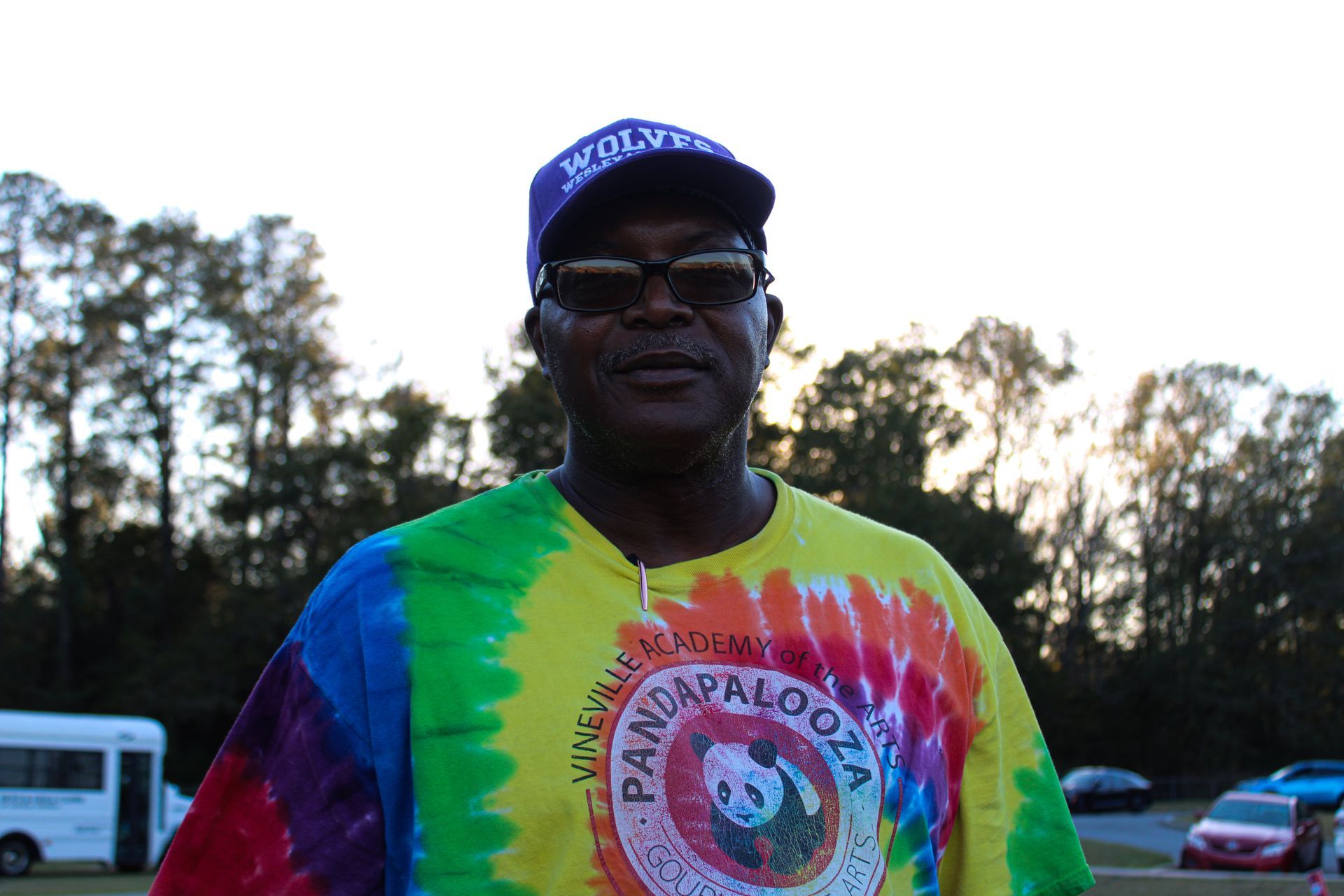 A man wearing a tie dye shirt and a hat stands in a parking lot