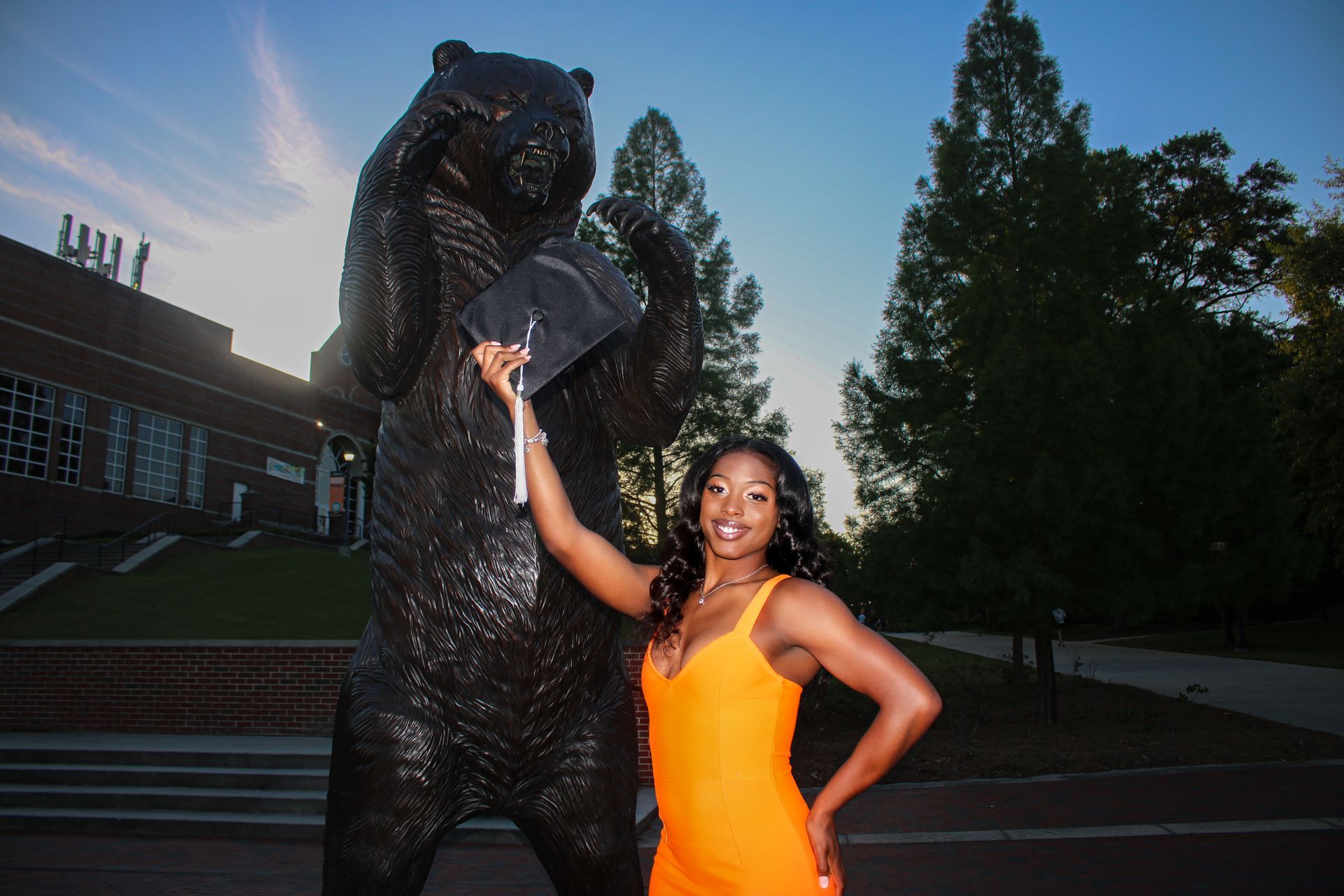 A woman in an orange dress is standing next to a statue of a bear holding a graduation cap.