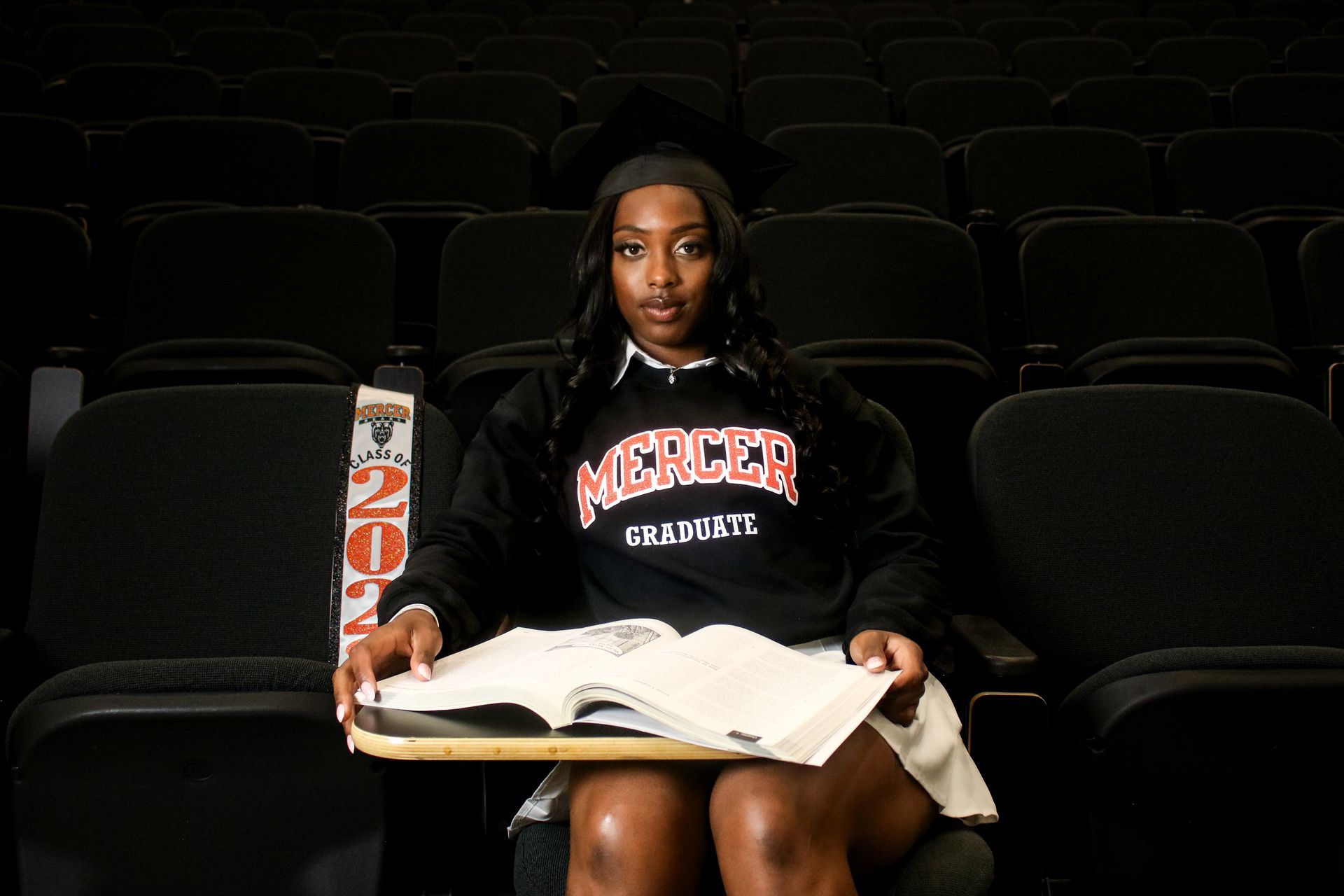 A woman in a cap and gown is sitting in an auditorium holding a book