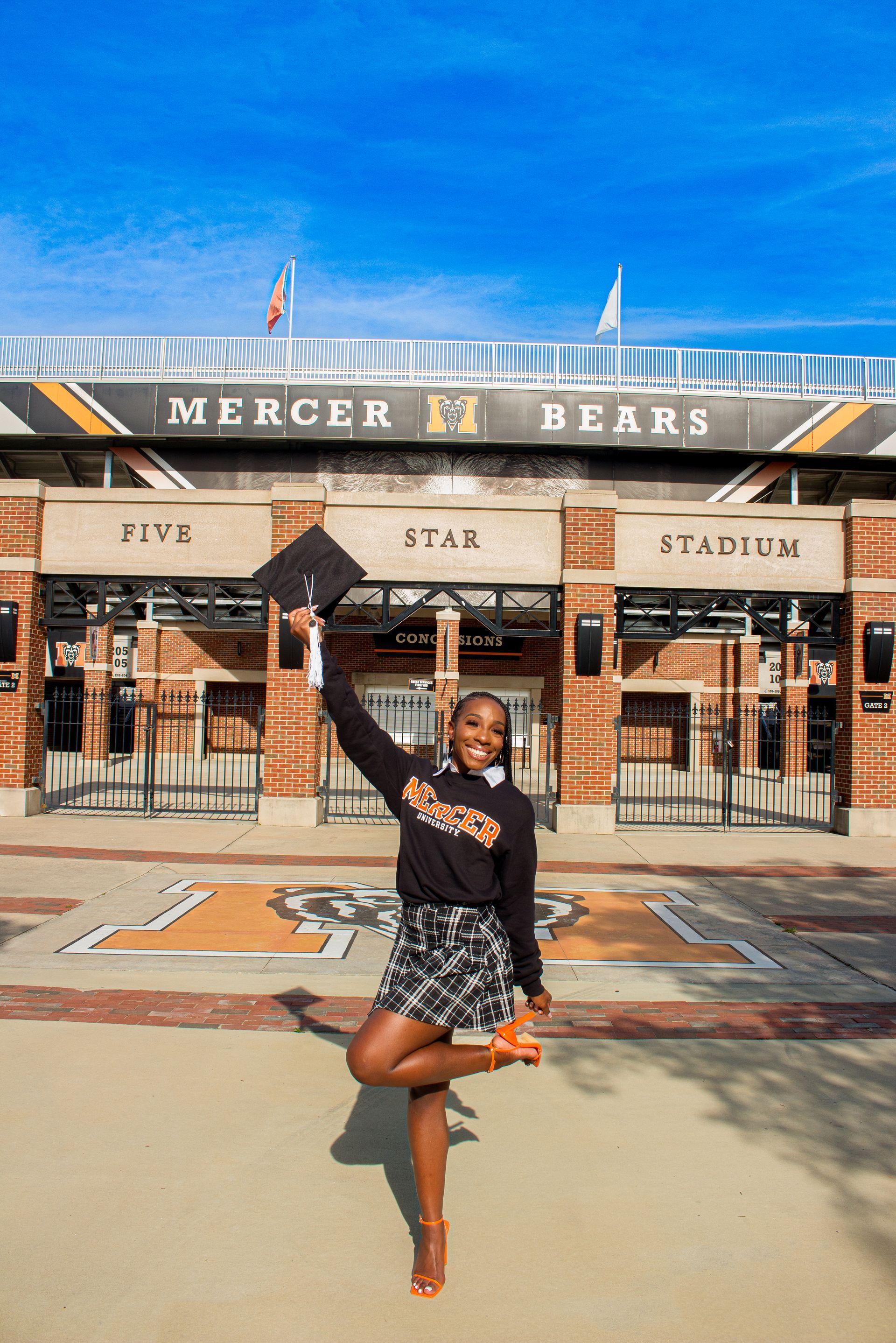 A woman wearing a graduation cap and gown is standing in front of a stadium.