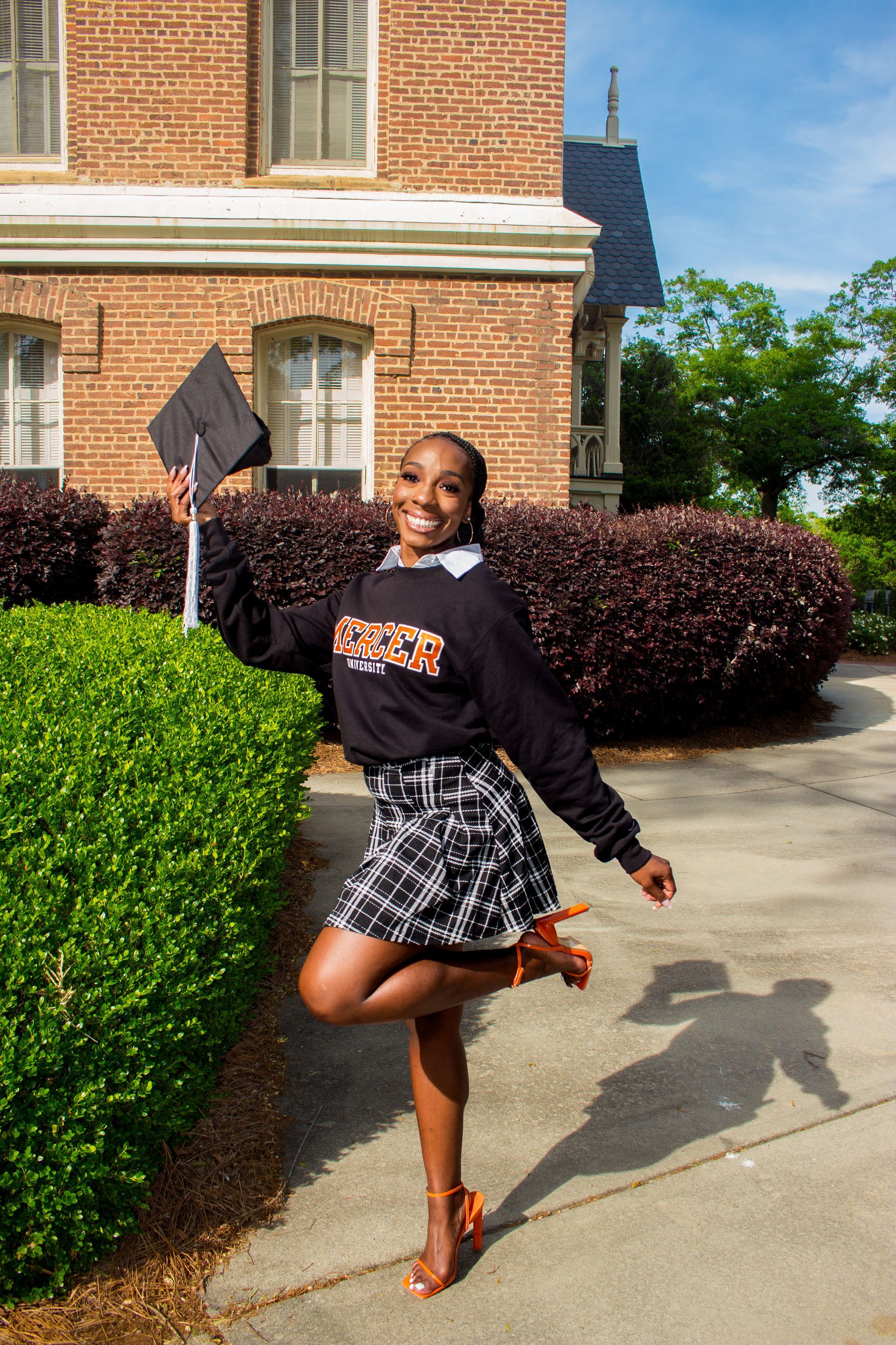 A woman is standing on one leg in front of a brick building holding a graduation cap.