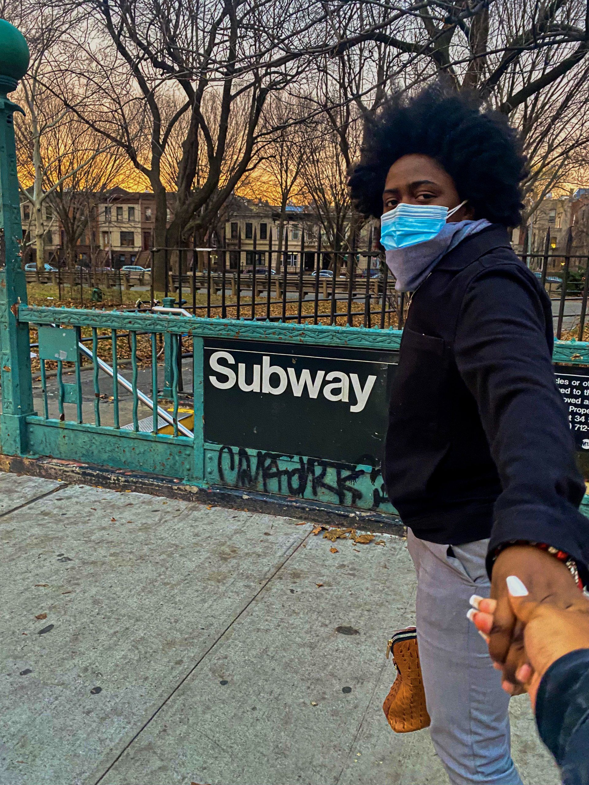 A woman wearing a mask is holding a man 's hand in front of a subway sign.