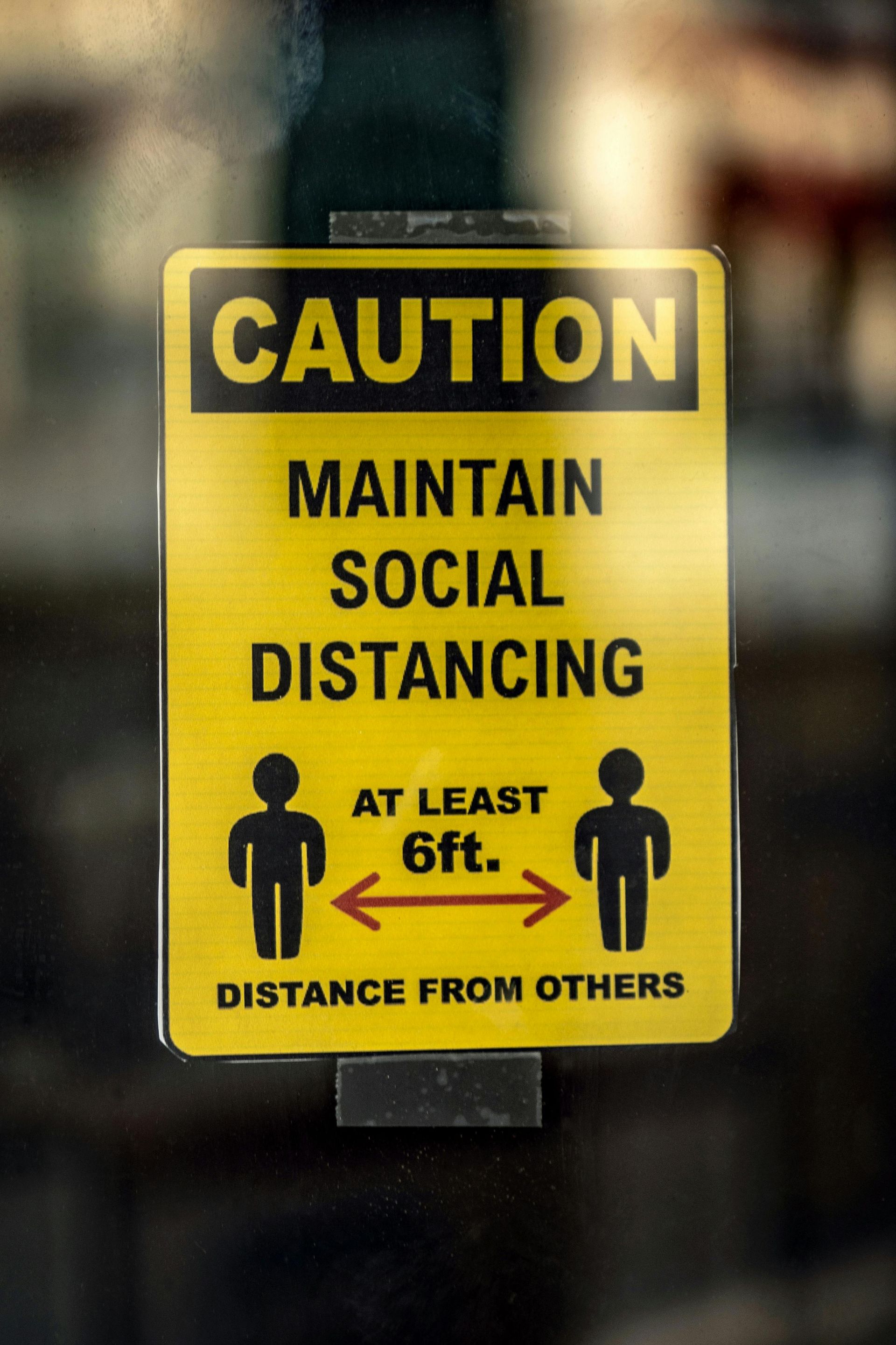 A yellow sign that says caution maintain social distancing at least 6 ft distance from others