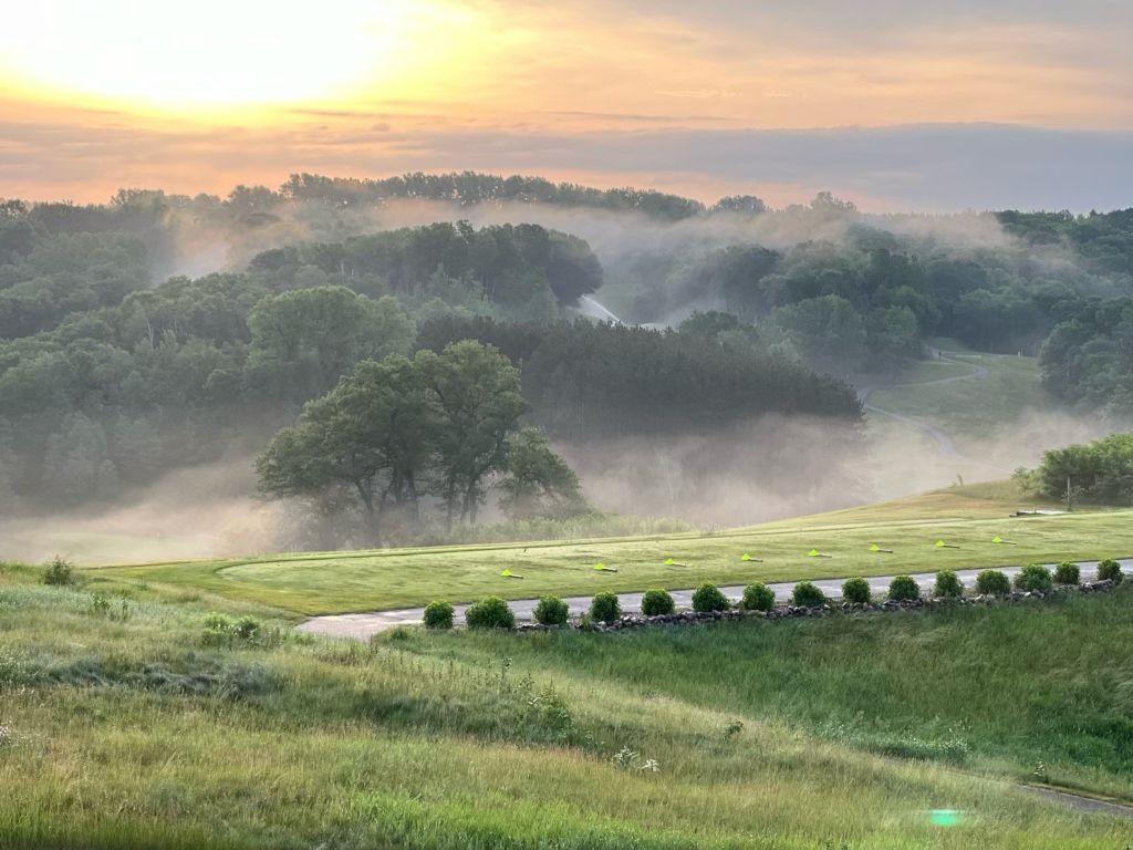 The sun is setting over a foggy field with trees in the background.