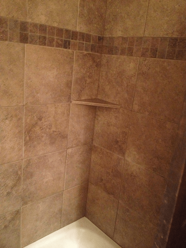 A shower with brown tiles and a shelf in the corner.