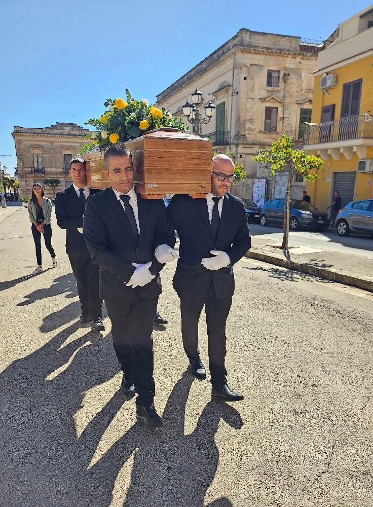 Un gruppo di uomini trasporta una bara di legno con dei fiori sopra.