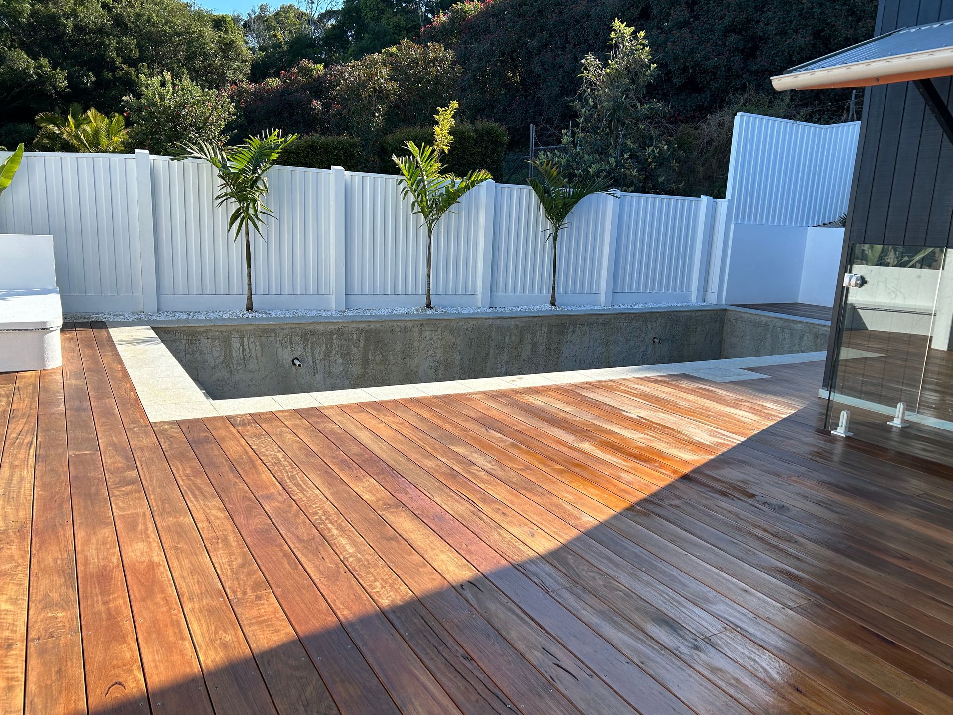 Wooden deck surrounds a rectangular concrete pool, white fence and trees in background — CM Timber Flooring in East Ballina, NSW