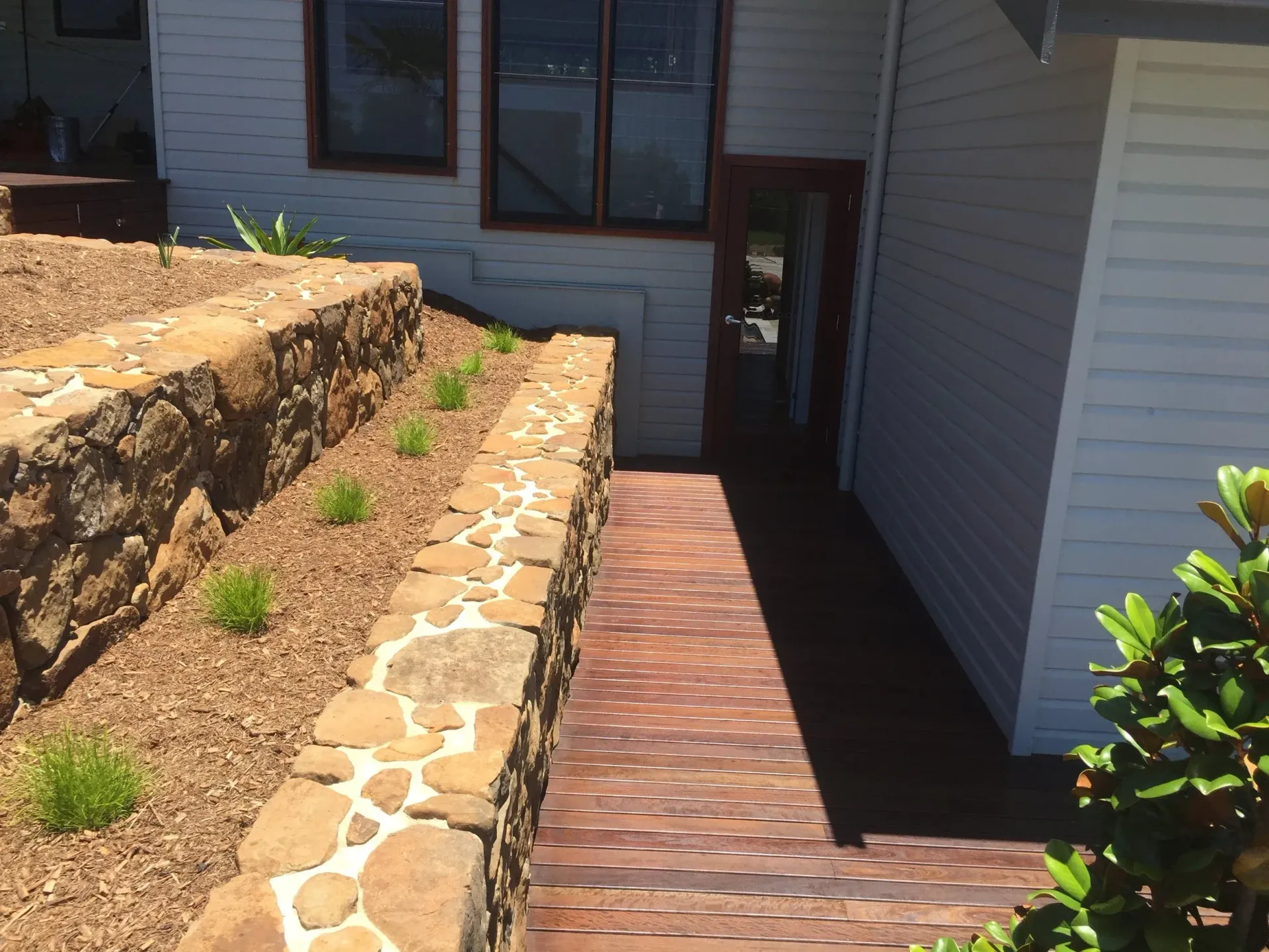 Stone retaining walls border a brick walkway leading to a doorway, with a white-sided house on the right — CM Timber Flooring in East Ballina, NSW