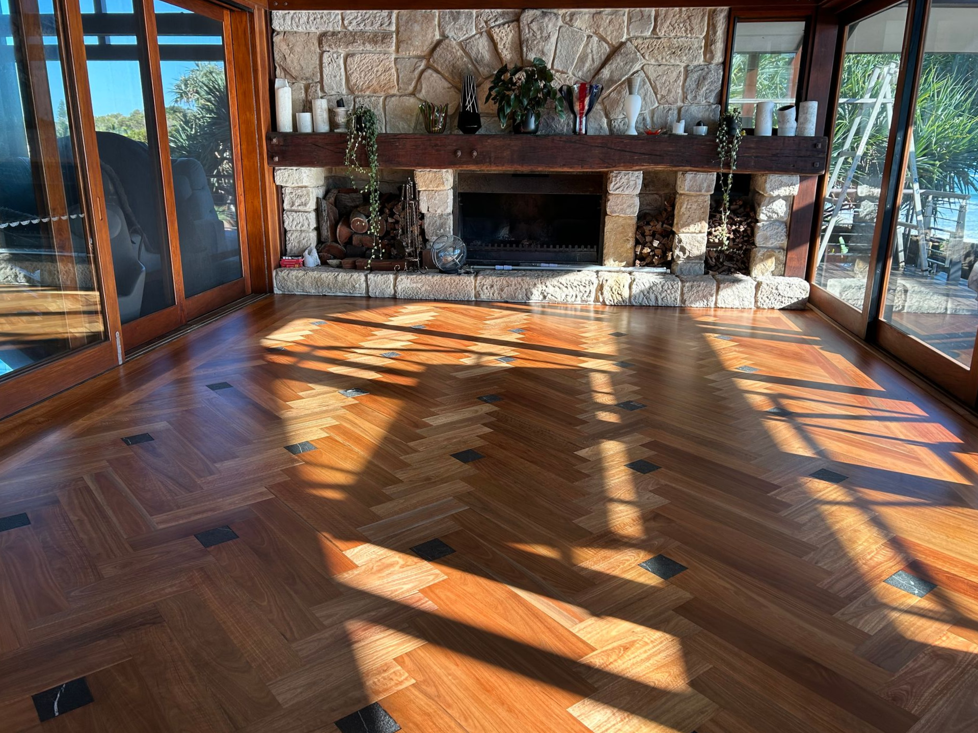 Wooden-floored room with stone fireplace, flanked by windows. Sunlight streams across the floor — CM Timber Flooring in East Ballina, NSW