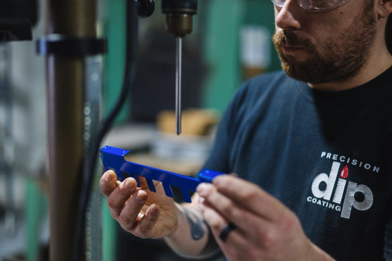 A worker in a Precision Dip Coating shirt inspects a blue, notched metal part under a drill press.