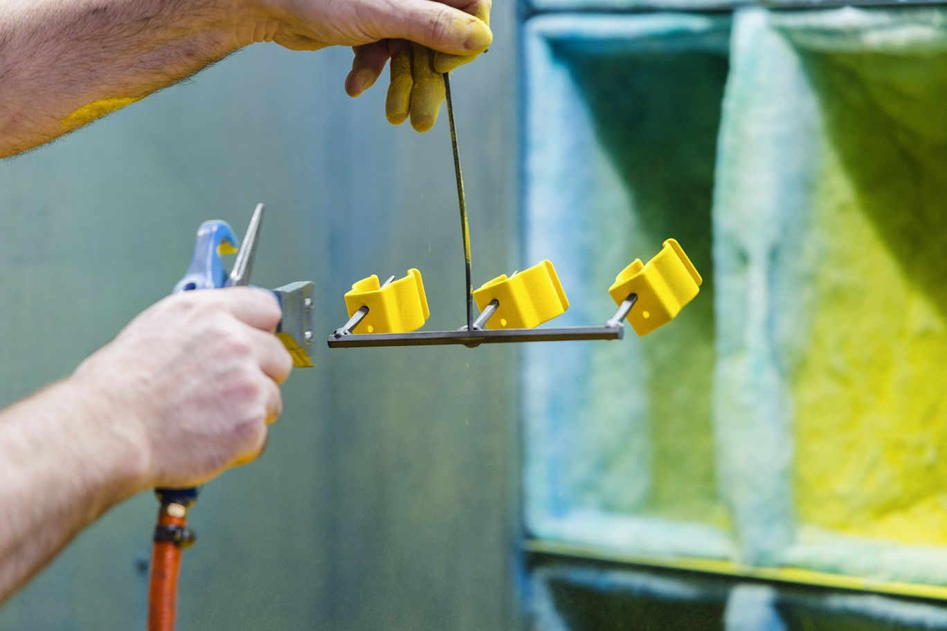 A worker sprays yellow powder coating onto small metal parts hanging from a wire in an industrial workshop.