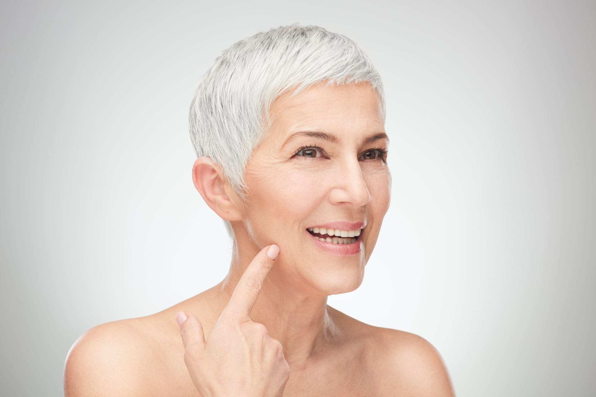 A woman with short gray hair is smiling and pointing at her face.