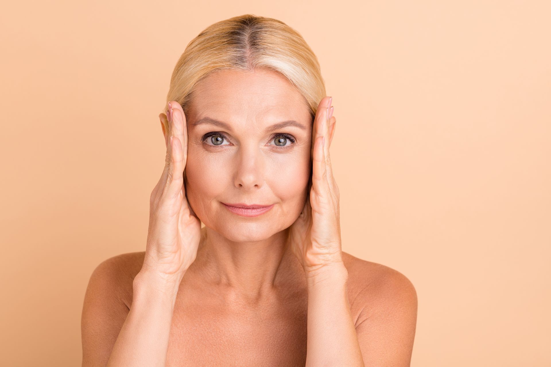 Woman with hands on her face, against a beige backdrop, looking directly at the viewer.