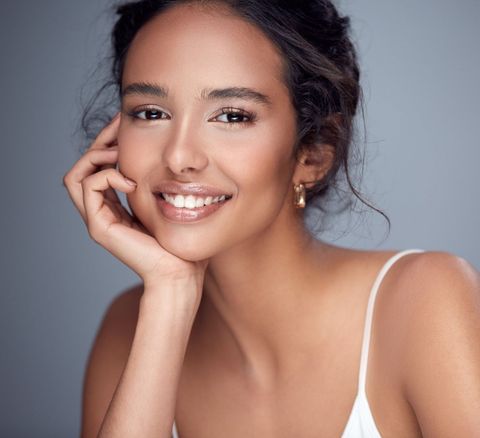 A woman in a white tank top is smiling with her hand on her face.