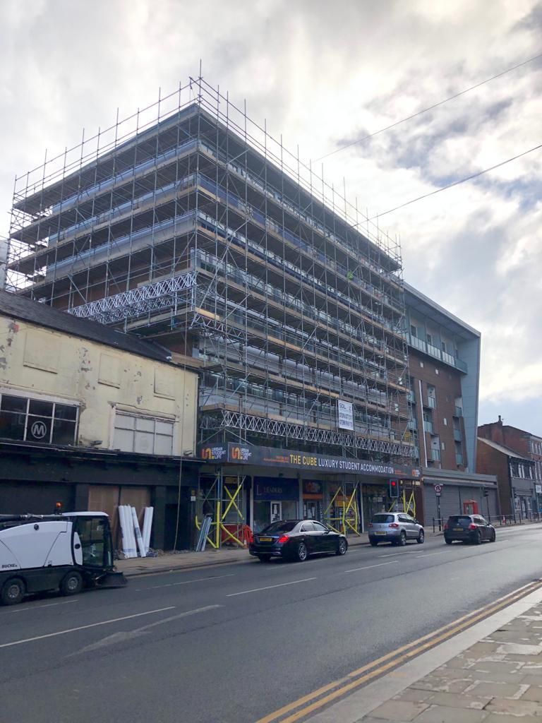 Multi-story building under construction, covered in scaffolding, street view. Cars parked along road.