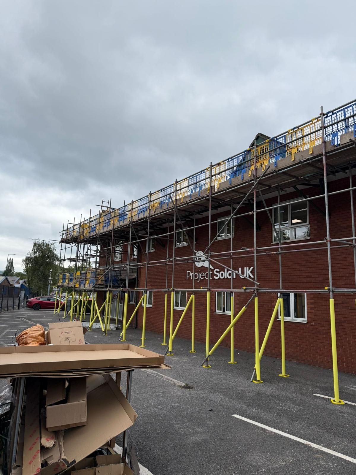 Building with scaffolding, brick exterior, cloudy sky. Cardboard debris in foreground. 