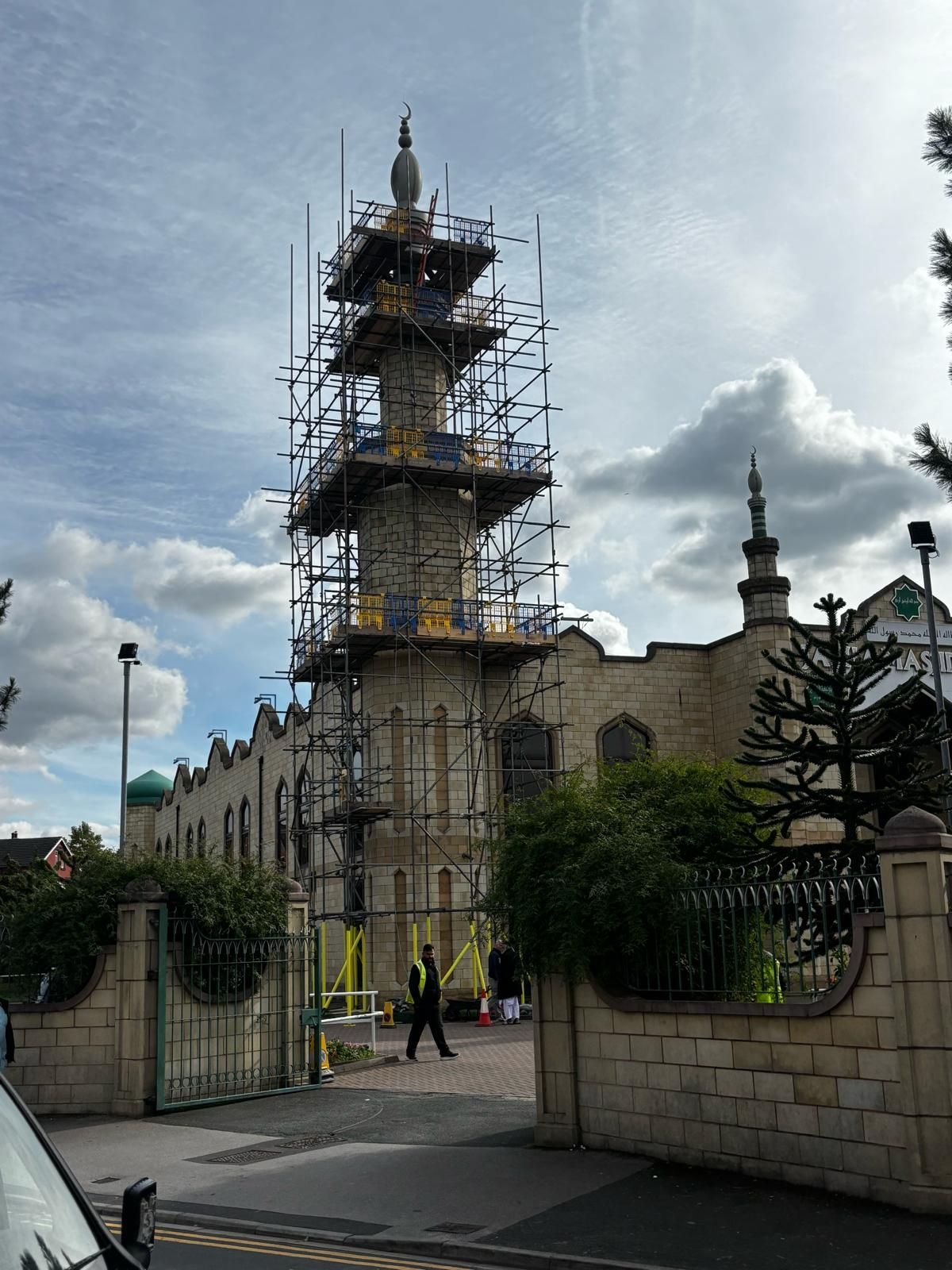 A mosque under construction with scaffolding on its minaret. A person walks in front. Cloudy sky.