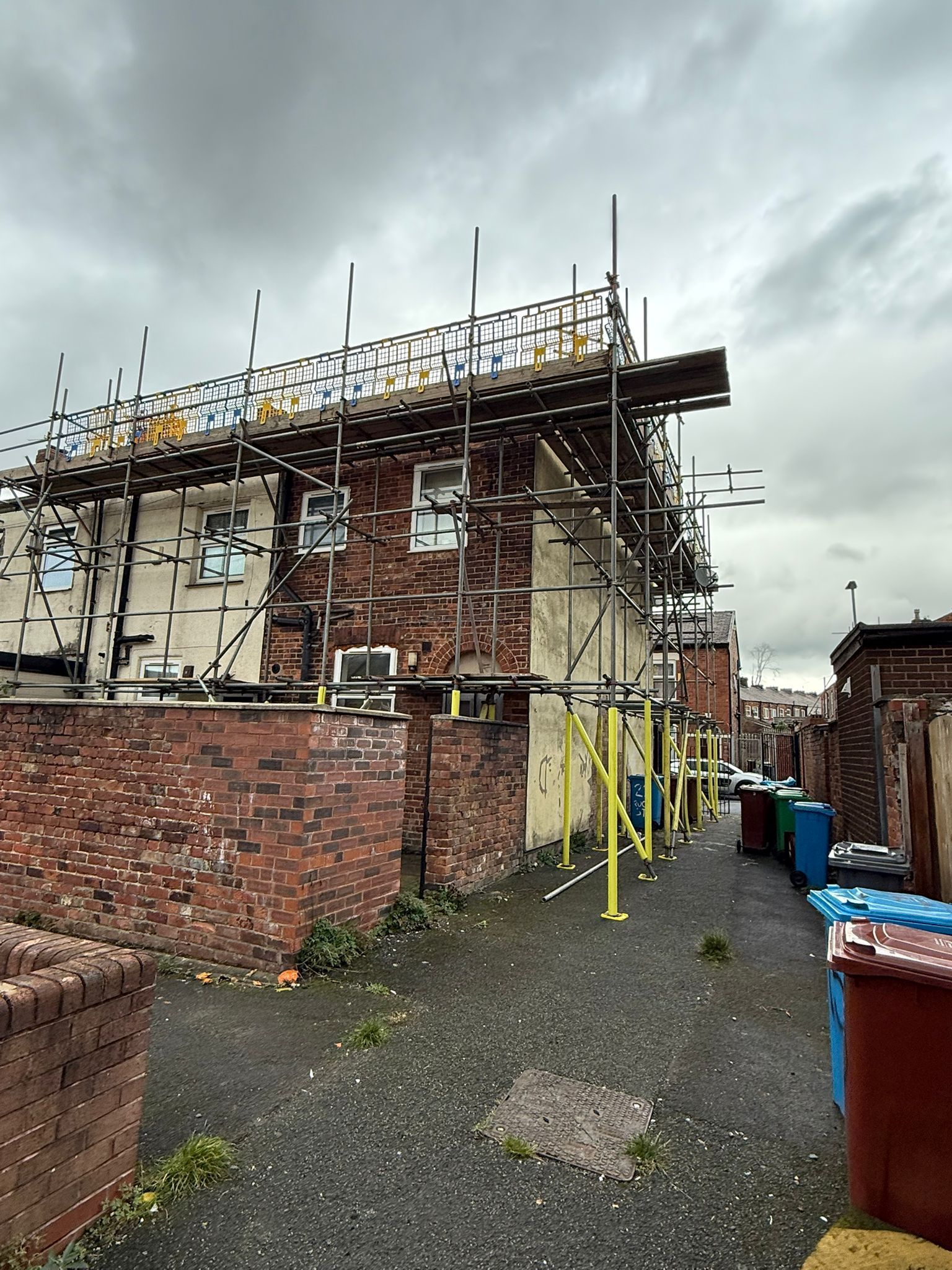 Building under construction, scaffolding wraps two-story brick building, overcast sky, alley with trash bins.