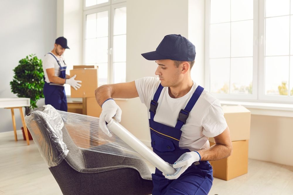Two movers wrapping a chair in plastic wrap; indoor, preparing for relocation.