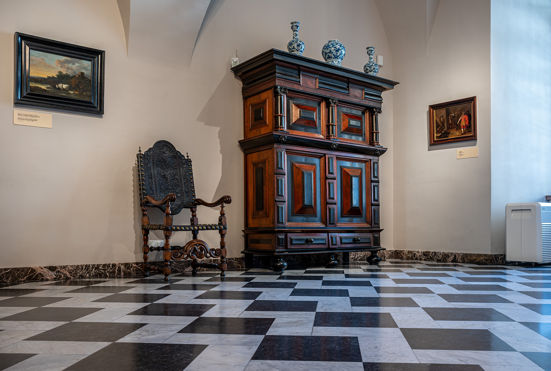 A museum room with a large antique cabinet, chair, paintings, and checkered floor.