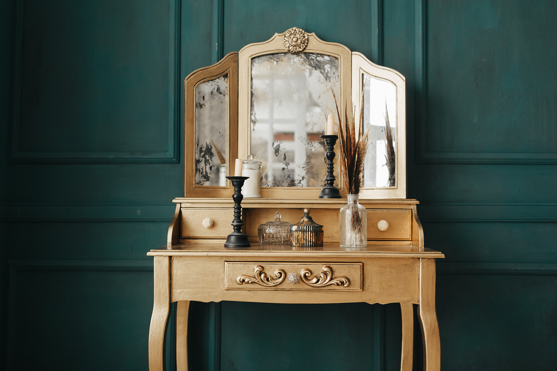 Gold vanity table with a tri-fold mirror and decorative items against a teal wall.