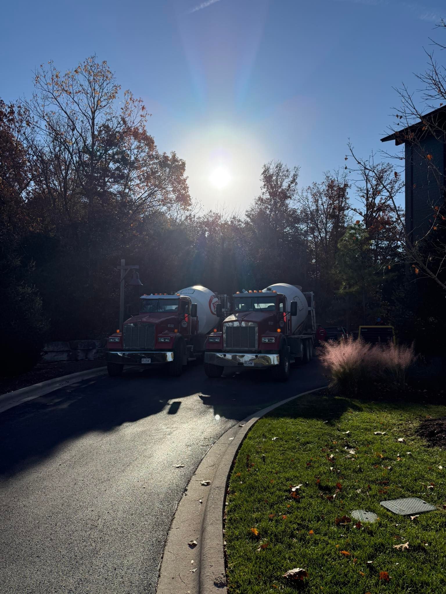 Two concrete mixer trucks parked on a driveway with the sun shining behind trees.
