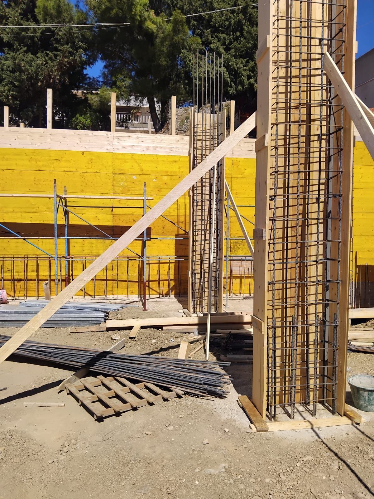 Cantiere con casseforme in legno e barre di armatura per pilastri in cemento, contro una parete in legno giallo.