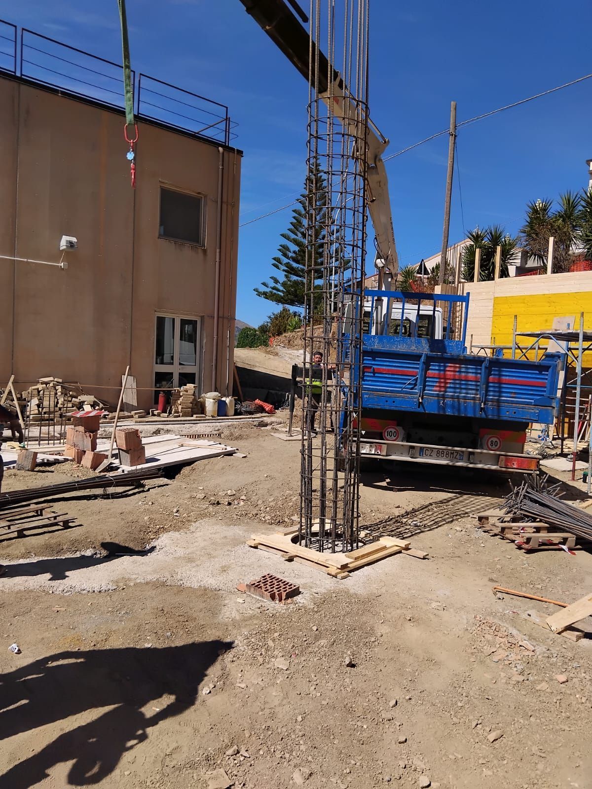 Cantiere con gabbia di tondini di acciaio posta su una base di cemento; camion blu, edificio beige, giornata di sole.
