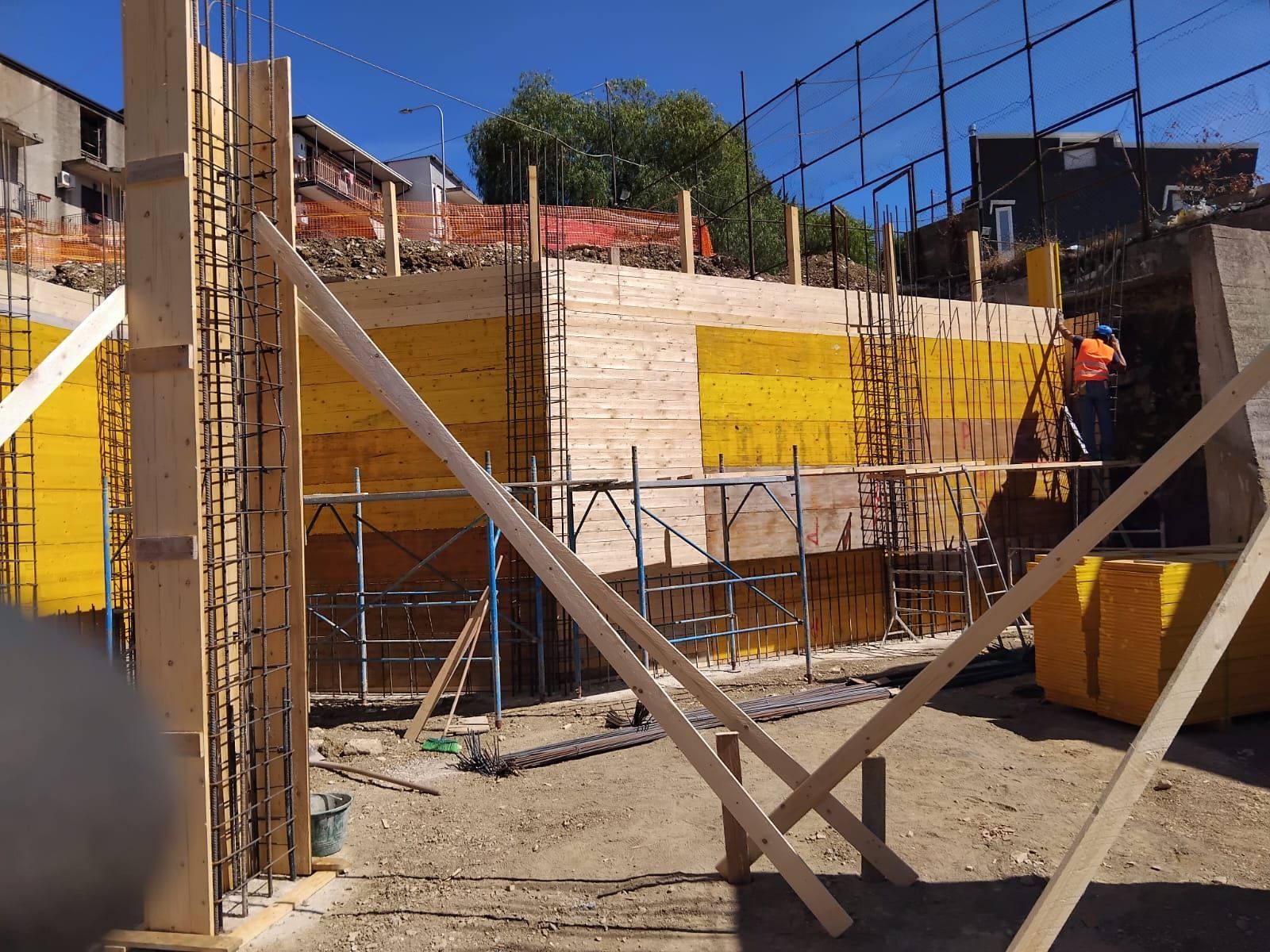 Cantiere con casseforme in legno e barre di armatura per muro di cemento, sostenuto da impalcature; luce solare intensa.