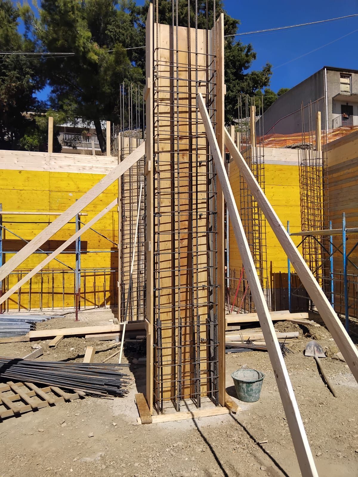 Cantiere: cassaforma in legno con barre di armatura, controventata, contro un muro giallo e un cielo azzurro.