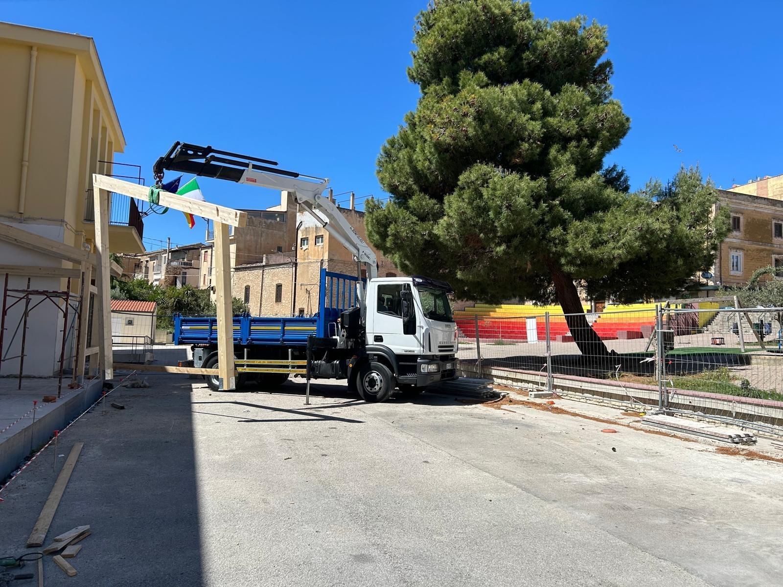 Un camion con una gru solleva travi di legno vicino a un edificio e a un grande albero in un ambiente esterno soleggiato.