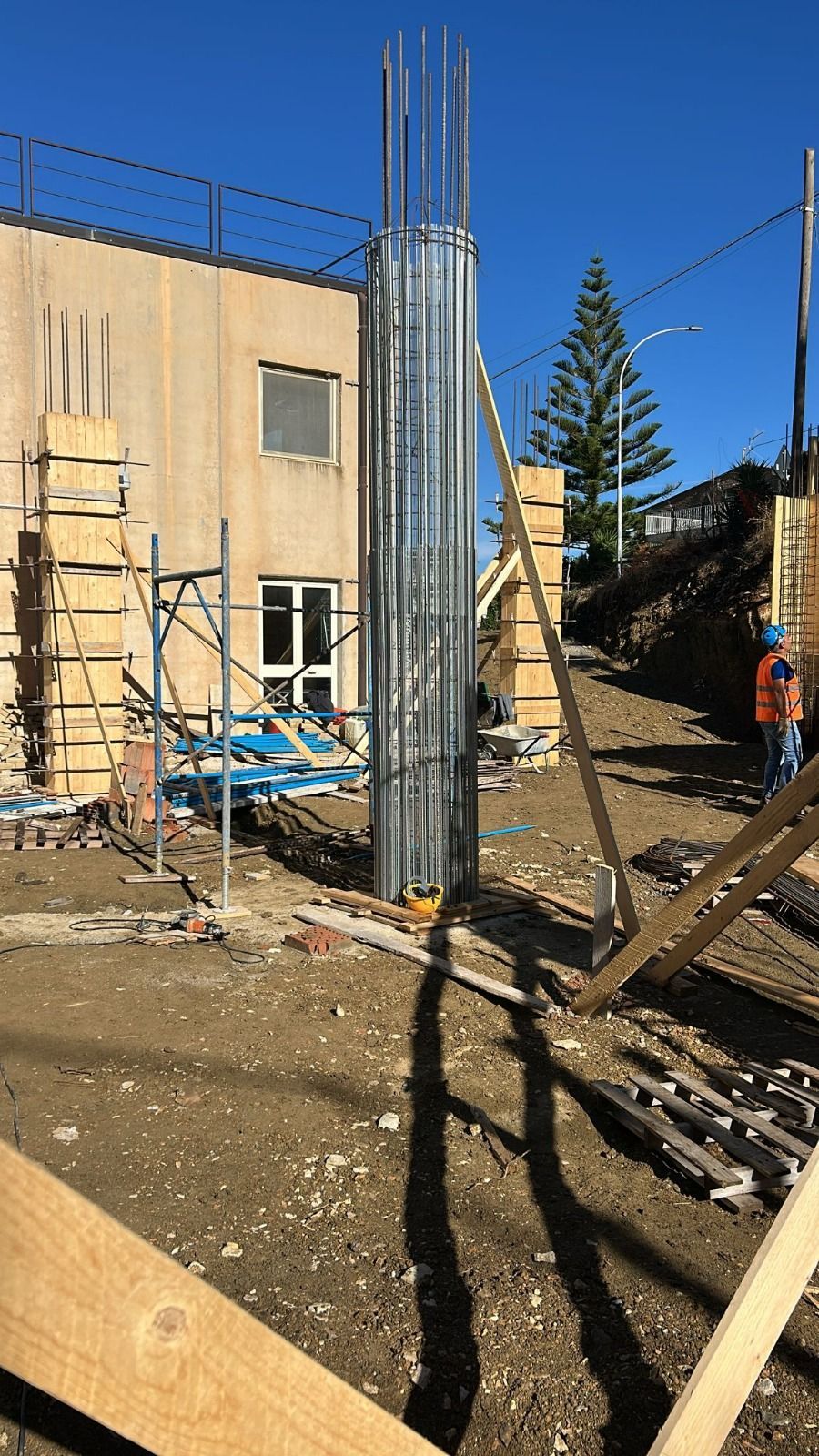 Cantiere con casseforme in metallo e legno per una colonna di cemento, accanto a un edificio in costruzione.