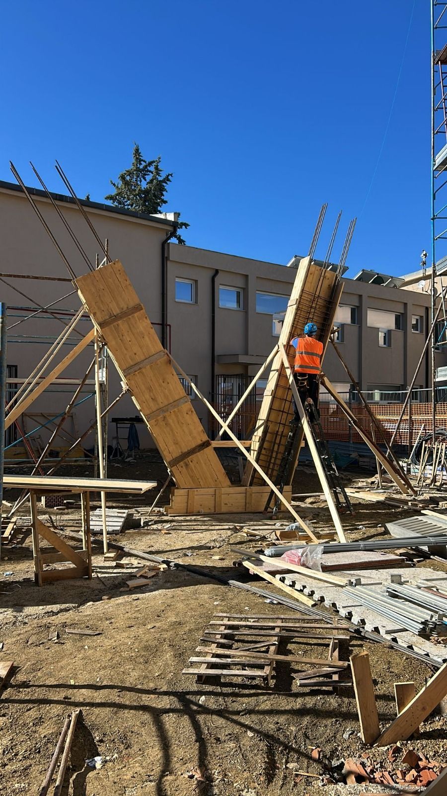 Cantiere con casseforme in legno, un operaio e un edificio sotto un cielo azzurro.