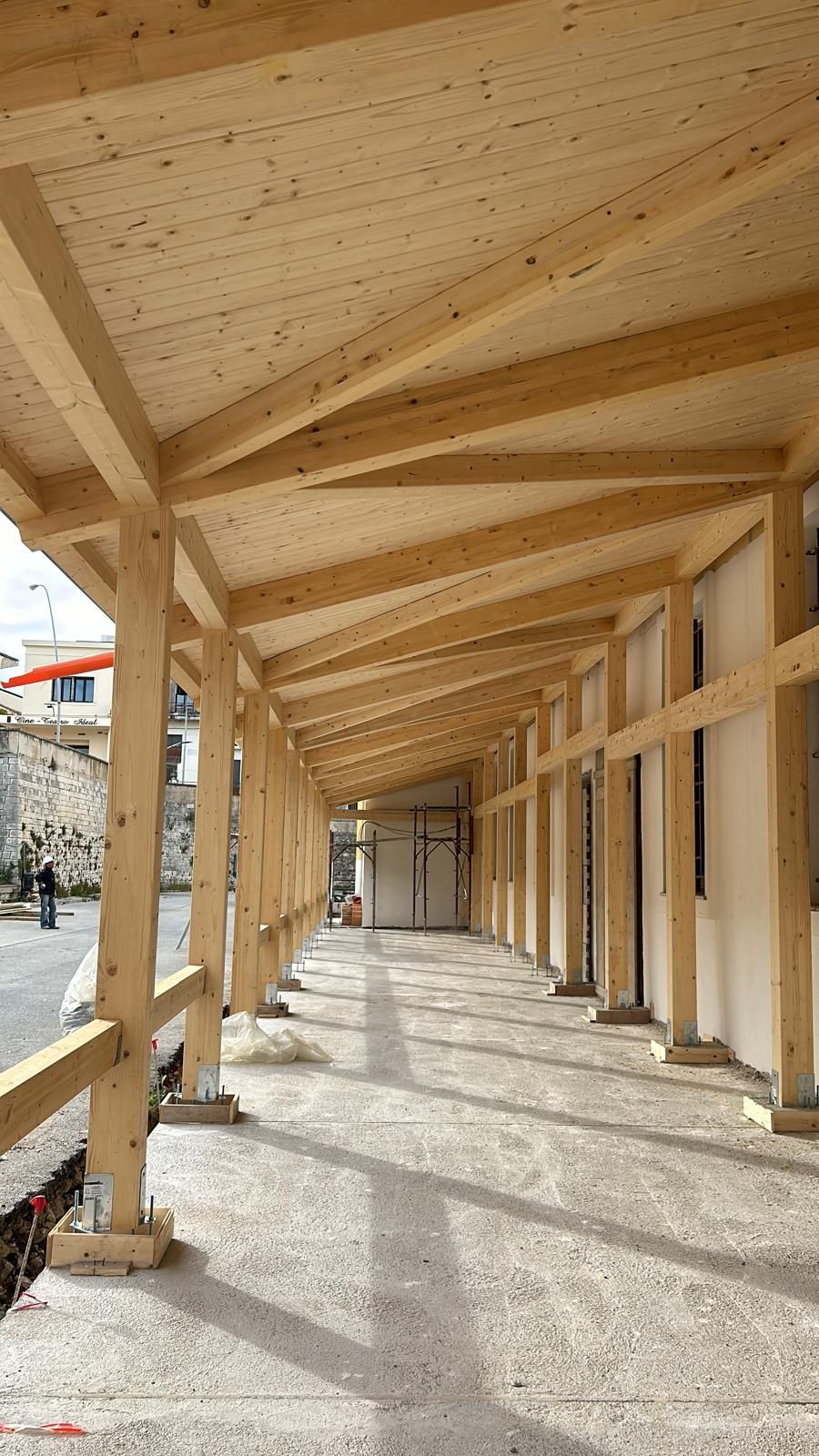 Passerella coperta in legno in uno spazio pubblico, sostenuta da pali. La luce del sole proietta lunghe ombre.